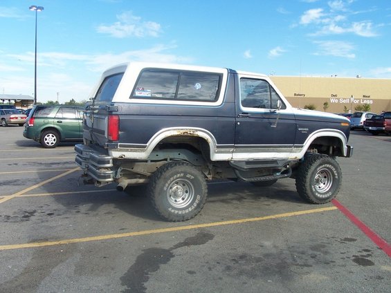 Lifted blue and white Ford Bronco parked in a lot, side view. Another SUV is parked behind it