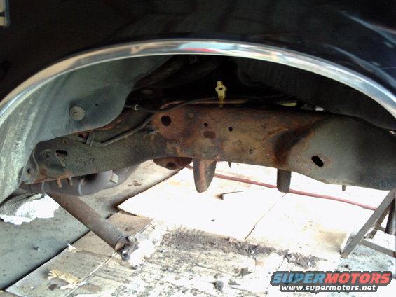 Black vehicle's rusty frame and suspension components, viewed from below and behind the wheel well