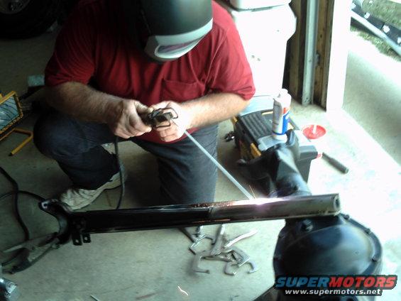 Welder in red shirt and welding mask works on a black metal frame part with sparks flying