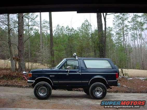 Dark blue Ford Bronco SUV, side view, parked on dirt road with trees in background