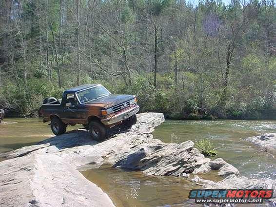 Dark blue and orange Ford Bronco on rocks near water, front-left view