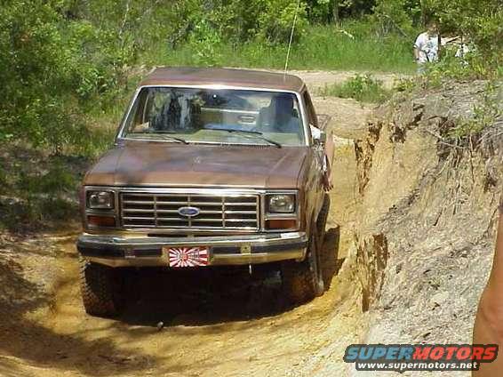 Brown Ford pickup truck driving on a dirt path with steep sides. It has a Japanese flag sticker on the front