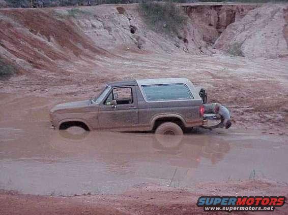 Ford Bronco stuck in mud pit, person bent over at rear bumper