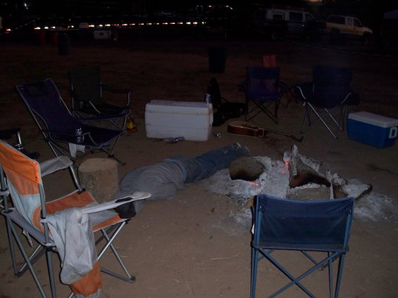 person lying by a campfire with chairs and coolers around