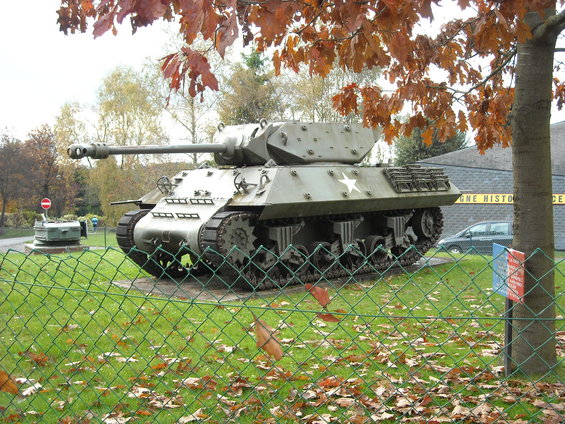 Sherman tank with a white star on its side, parked outside a building with text that reads 'GNE HISTO CE'