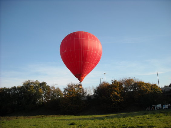Red hot air balloon in a blue sky over a grassy field and trees