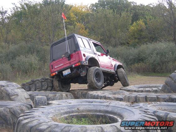Red Suzuki Samurai driving over large tires on an off-road course
