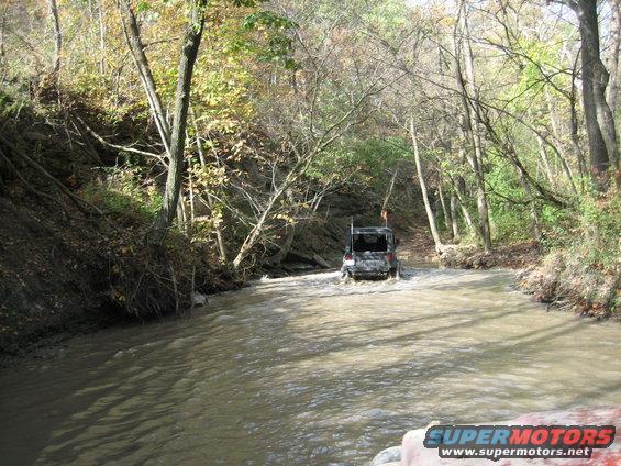 Black Jeep Wrangler driving through a muddy river surrounded by trees