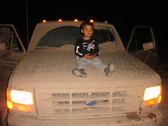 Kid in skeleton shirt sits on the hood of a dusty Ford truck. Another truck is visible to the left