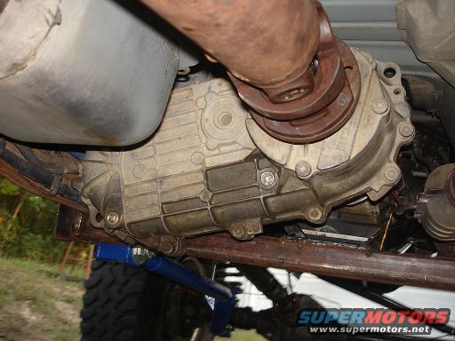 Underside view of a vehicle's transfer case and driveshaft assembly. A blue jack is visible below