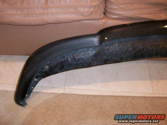 Carbon fiber car bumper part with visible mounting holes and textured surface, resting on a floor with a leather couch in the background