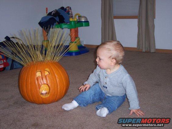 Jack-o-lantern with straw hair and a lit candle inside sits on carpet next to a baby in jeans and a grey shirt
