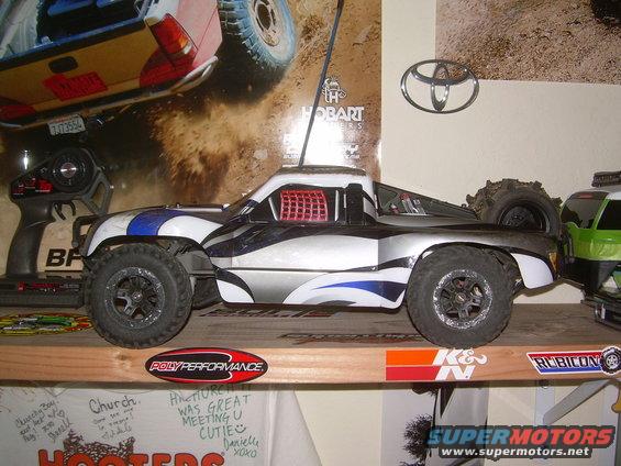 Remote control short course truck with blue and silver graphics, next to a remote control, and a Toyota logo on the wall