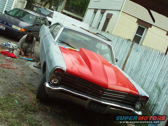 1967 Ford Galaxie with red hood scoop, parked outside next to another car. Person is working on something on the ground