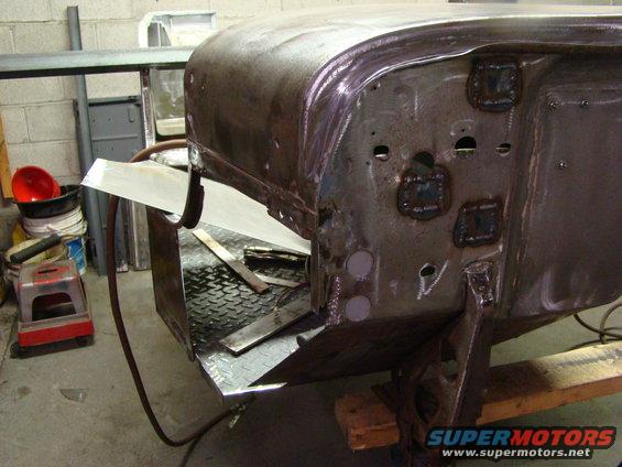 Unfinished vehicle body panel with visible welds and cutouts, resting on a wooden support