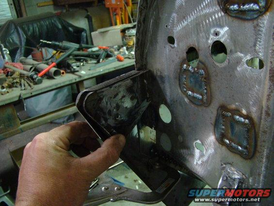 Hand holding a bent metal bracket against a larger metal panel with numerous welds and circular holes