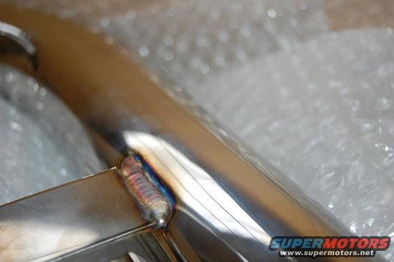 Close-up of a shiny metal pipe with a weld seam, wrapped in bubble wrap. Supermotors.net logo visible