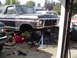 1979 Ford Bronco - Mechanics work on a black Ford Bronco lifted on jack stands in a driveway, with another truck visible in the background.
