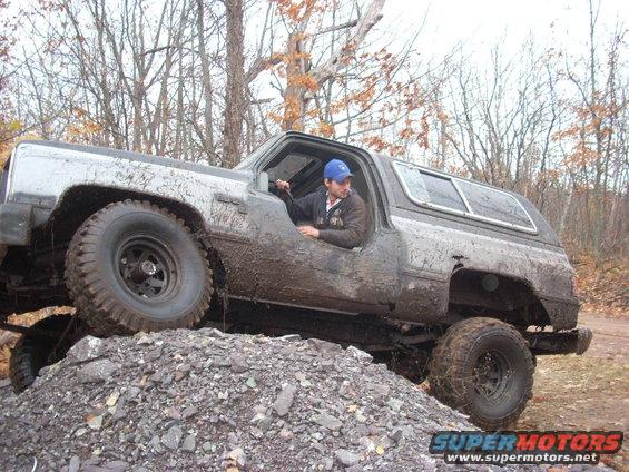 Muddy SUV climbing a pile of rocks with a driver inside. The vehicle appears to be a Chevrolet K5 Blazer from the late 1980s