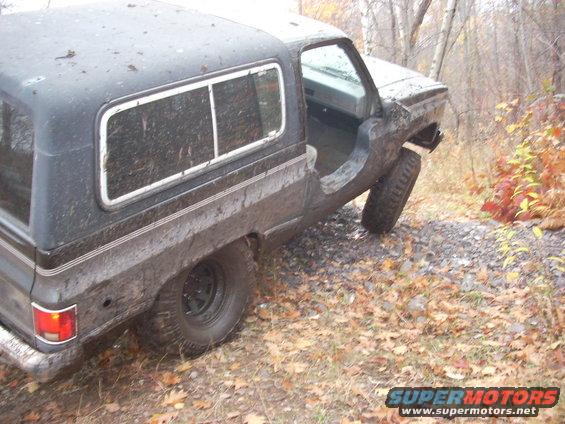 Dark SUV with camper shell tilted on a gravel path, driver's door open