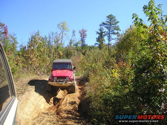 Red Jeep Wrangler on a dirt trail, front bumper visible, with another vehicle's side in the foreground