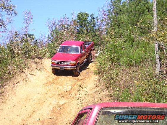 Red pickup truck driving up a dirt hill, another red vehicle in foreground