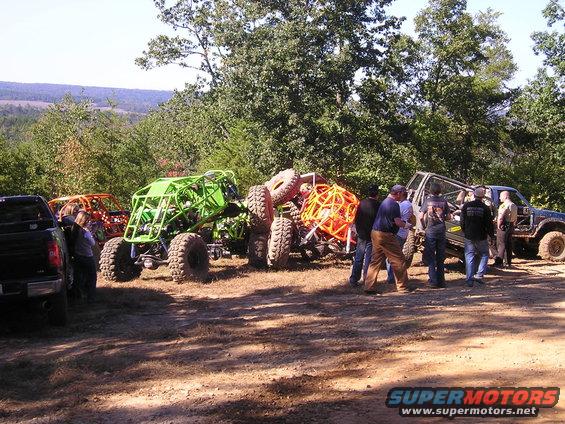 Several custom off-road buggies and a dark pickup truck parked on a dirt lot with trees in the background