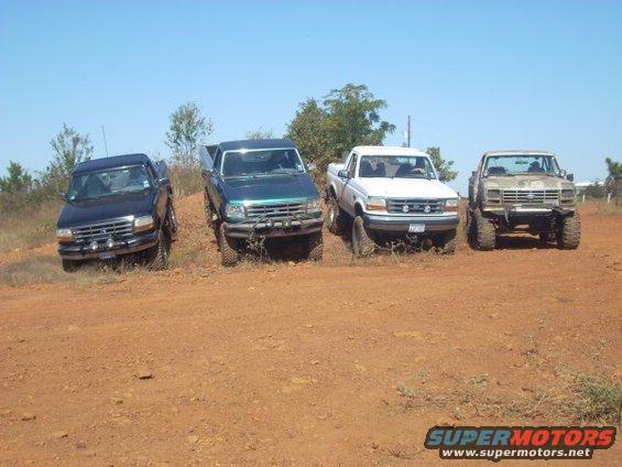 Four pickup trucks, including a 1992 Ford Bronco, parked on a dirt hill under a clear blue sky