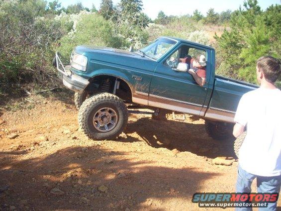 Green Ford F-series pickup truck with lifted suspension and off-road tires driving on a dirt trail