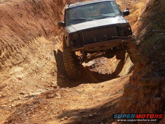 Off-road vehicle with lifted suspension driving through a narrow, deep dirt rut. Front grille is missing