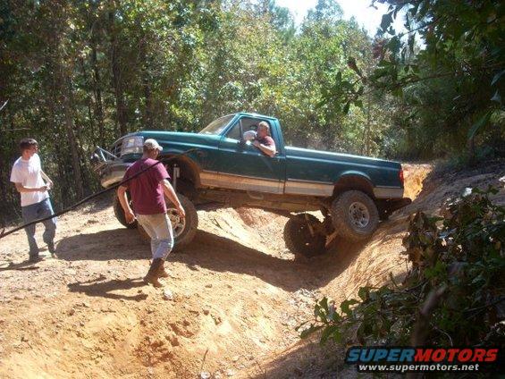 Green pickup truck with lifted suspension navigating a steep dirt incline, two people outside the vehicle