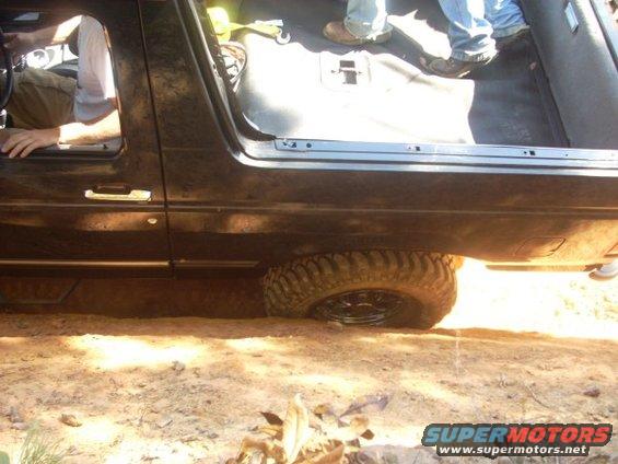 Black SUV with off-road tires stuck in dirt, side view. Interior visible with people