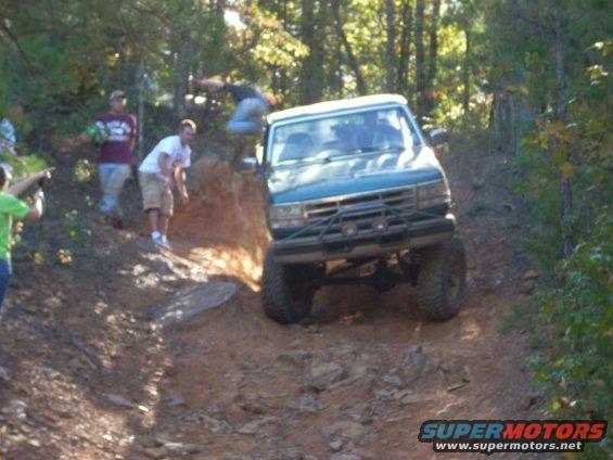 Green SUV with a brush guard going up a rocky trail, people watching