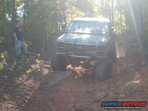 Ford Bronco with large tires navigating a muddy trail, person standing nearby