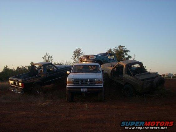 Four trucks and SUVs parked on dirt, including a black pickup, a white Ford Ranger, a teal Ford F-Series, and a tan Bronco. The white truck is in the foreground