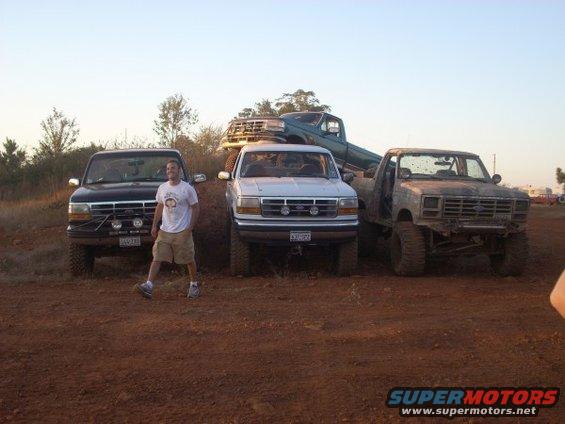 Man standing next to a black truck, with a white truck and a green truck behind it. A fourth, older truck is on the right