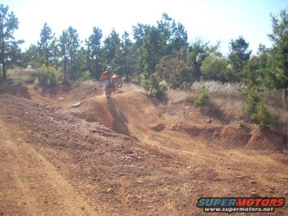 Dirt bike rider on a hill, orange shirt, blue jeans, dirt path, trees in background