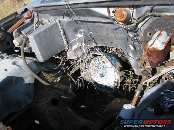 Engine bay of a weathered truck, showing rusted components, wiring, and debris. A blue-domed headlight is visible