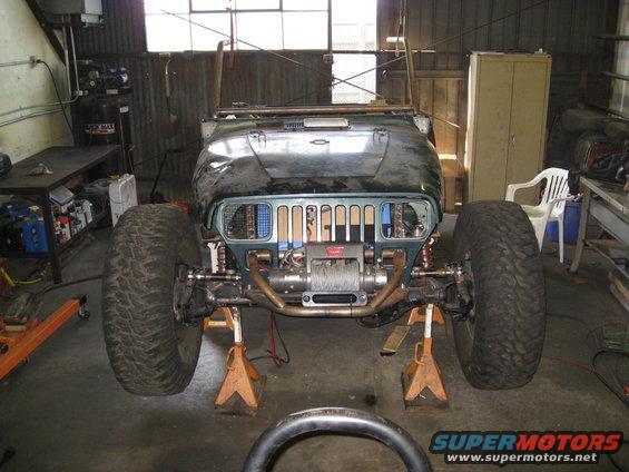 Modified Jeep on jack stands, front view. Dark green body, large tires, winch installed