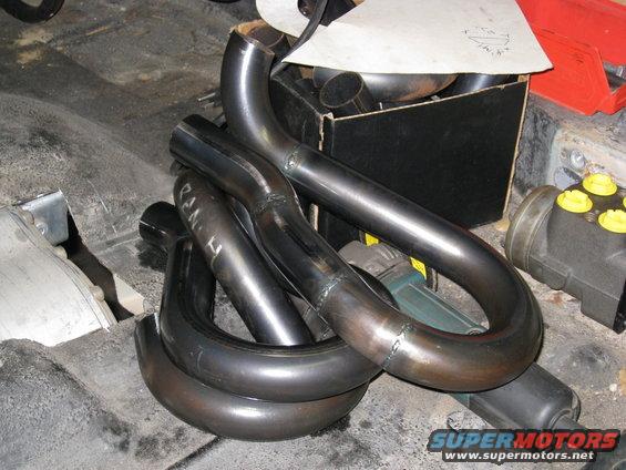 Pile of bent metal exhaust pipes and a black box with more pipes inside, on a floor with some tools