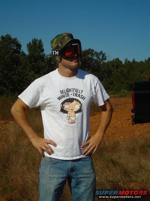 Man wearing a camo hat, goggles, and a white t-shirt with a cartoon character and text. He's standing outdoors with trees and a blue sky