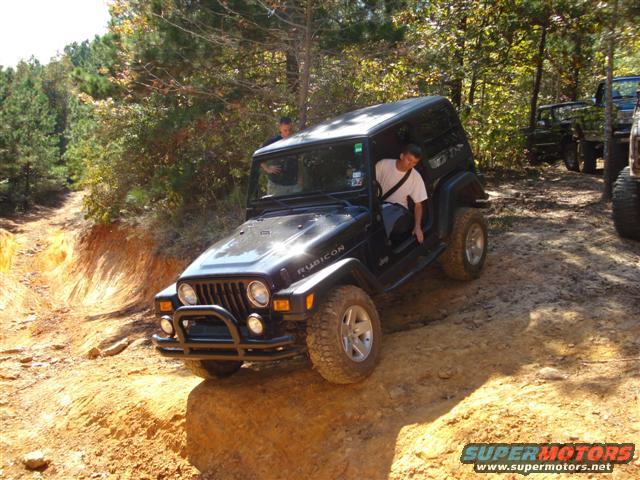 Black Jeep Wrangler Rubicon with a man in the driver's seat, driving on a dirt trail with trees in the background