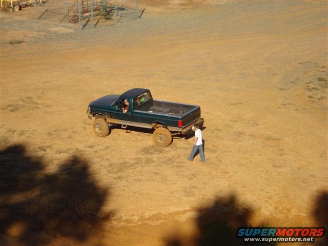 Green pickup truck driving on a dirt field with a person walking nearby
