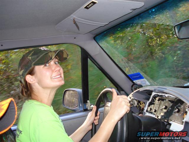 Woman wearing a camo hat and green shirt driving with the dashboard partially removed. Trees are visible through the windshield