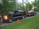 1998 Chevrolet Tahoe - A black Chevrolet Suburban from the mid-1990s is seen towing a trailer loaded with a skid steer loader on a gravel road.