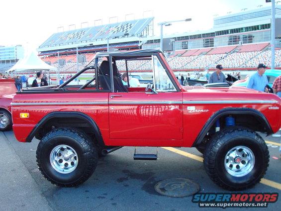 Red 1977 Ford Bronco with a black soft top and roll cage, seen from the side