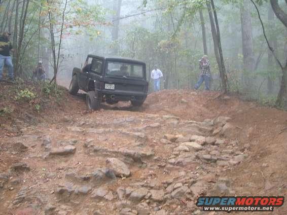 Black SUV driving up a rocky, wooded trail. People are watching