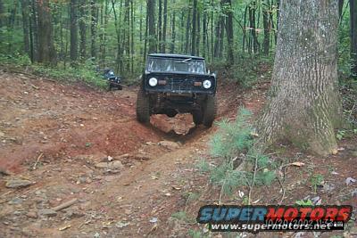 1972 Ford Bronco driving on a muddy forest trail; another vehicle is visible in the background