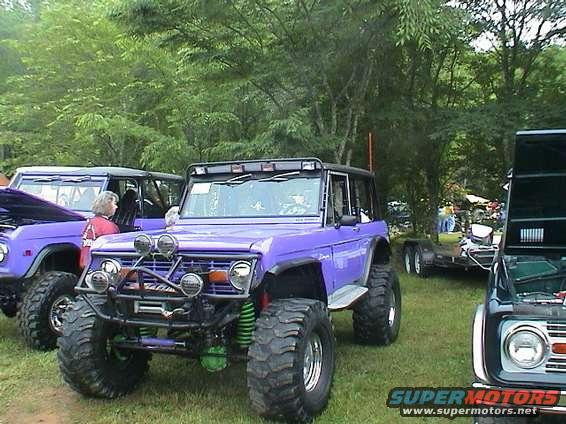 1972 Ford Bronco, purple, lifted with large tires and off-road lights. Another Bronco and a trailer are visible