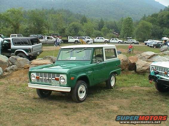 Green 1970 Ford Bronco with a white hardtop, parked outdoors on a grassy area with other vehicles in the background